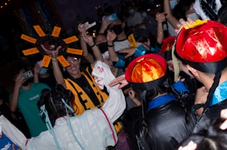 A celebratory crowd is gathered with individuals wearing colorful costumes and hats. Some participants are holding up phones to capture the event. Distinctive elements include a large decorative sun-shaped object with text panels, and a woman in a white outfit with long braids and blue ribbons.