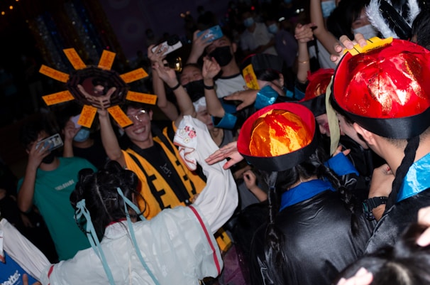 A celebratory crowd is gathered with individuals wearing colorful costumes and hats. Some participants are holding up phones to capture the event. Distinctive elements include a large decorative sun-shaped object with text panels, and a woman in a white outfit with long braids and blue ribbons.