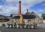 A distillery courtyard with a prominent display featuring black barrels, each marked with a letter to spell 'BALBLAIR.' A large copper distillation apparatus and a tall red chimney stand in front of a traditional stone building labeled 'reception.' Wooden benches and planters with flowers add to the scene, and stacked barrels are visible on the left. The background includes hills and a partly cloudy sky.