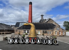 A distillery courtyard with a prominent display featuring black barrels, each marked with a letter to spell 'BALBLAIR.' A large copper distillation apparatus and a tall red chimney stand in front of a traditional stone building labeled 'reception.' Wooden benches and planters with flowers add to the scene, and stacked barrels are visible on the left. The background includes hills and a partly cloudy sky.
