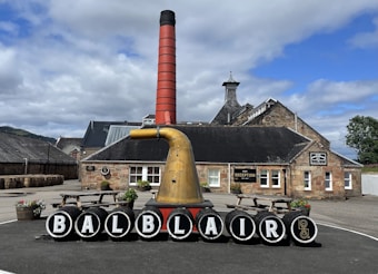 A distillery courtyard with a prominent display featuring black barrels, each marked with a letter to spell 'BALBLAIR.' A large copper distillation apparatus and a tall red chimney stand in front of a traditional stone building labeled 'reception.' Wooden benches and planters with flowers add to the scene, and stacked barrels are visible on the left. The background includes hills and a partly cloudy sky.