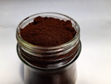 A glass jar filled with finely ground dark brown powder, likely coffee, placed on a plain surface. The texture of the powder is slightly uneven, with some clumps visible near the top. The jar has a clear, reflective surface that captures some ambient light.