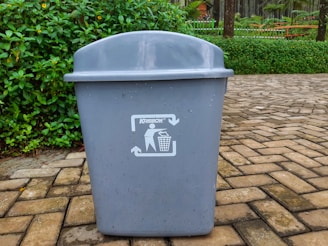 A large gray trash bin is prominently placed on a paved area with a backdrop of greenery and some flowering plants. The bin has a recycling symbol on it, accompanied by the word 'Krisbow'. The ground is made of interlocking stone tiles.