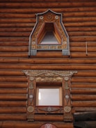 Two ornately decorated wooden windows are set within a log cabin wall. The upper window features triangular framing with intricate floral carvings and painted details in blue and gold. The lower window is rectangular with elaborate designs, including decorative elements of floral and geometric patterns, featuring similar color accents. The logs of the cabin are rich brown with visible texture.