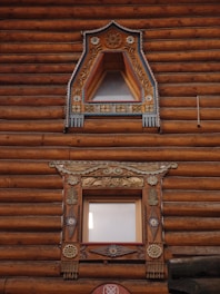 Two ornately decorated wooden windows are set within a log cabin wall. The upper window features triangular framing with intricate floral carvings and painted details in blue and gold. The lower window is rectangular with elaborate designs, including decorative elements of floral and geometric patterns, featuring similar color accents. The logs of the cabin are rich brown with visible texture.
