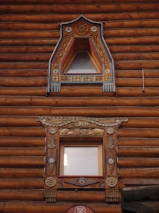 Two ornately decorated wooden windows are set within a log cabin wall. The upper window features triangular framing with intricate floral carvings and painted details in blue and gold. The lower window is rectangular with elaborate designs, including decorative elements of floral and geometric patterns, featuring similar color accents. The logs of the cabin are rich brown with visible texture.