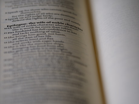 A close-up view of an open book with text discussing the qualities of a wife of noble character. The focus is on a specific passage, while the rest of the text is blurred, creating a depth of field effect.
