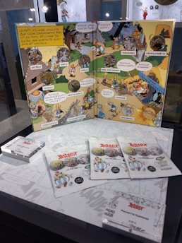 A comic book display featuring Asterix characters with coins integrated into the pages. The display includes several comic panels with speech bubbles and vibrant illustrations. Below the book, there are packets labeled 'Asterix' featuring characters from the comics, with pricing information shown.