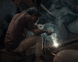 A person is engaged in welding, surrounded by sparks and smoke. The individual is bent over, focusing intently on the work. The setting appears dimly lit, creating a stark contrast with the bright welding sparks. The environment seems industrial, with metal objects in the background.