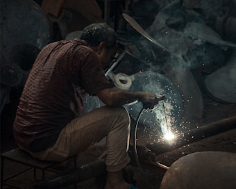 A person is engaged in welding, surrounded by sparks and smoke. The individual is bent over, focusing intently on the work. The setting appears dimly lit, creating a stark contrast with the bright welding sparks. The environment seems industrial, with metal objects in the background.