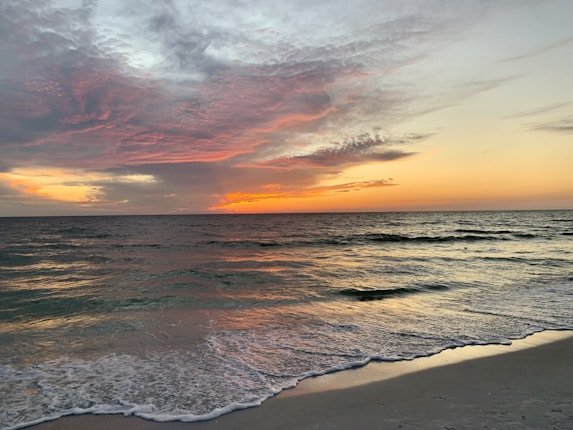 A serene beach setting with gentle waves lapping onto the shore. The sky is painted with stunning hues of orange and pink as the sun sets on the horizon, casting a warm glow over the ocean.