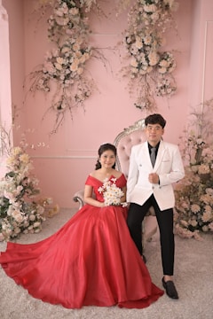 A couple is elegantly posed in front of an ornate floral backdrop indoors. The woman is seated on a vintage-style chair wearing a flowing red gown, holding a bouquet of flowers. Beside her stands a man dressed in a sharply tailored white jacket and black pants. The background features pale pink walls adorned with cascading floral arrangements featuring white and cream-colored blooms.