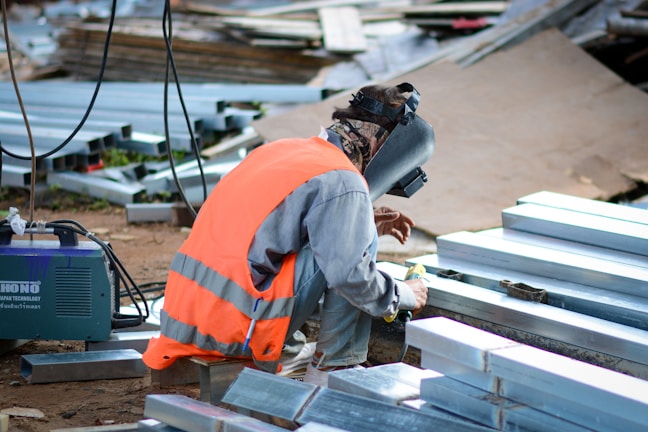 A person wearing an orange safety vest and a welding helmet crouches on the ground, working with metal beams. Nearby is a welding machine and various scattered metal pieces.