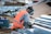 A person wearing an orange safety vest and a welding helmet crouches on the ground, working with metal beams. Nearby is a welding machine and various scattered metal pieces.