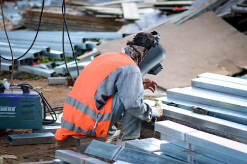 A person wearing an orange safety vest and a welding helmet crouches on the ground, working with metal beams. Nearby is a welding machine and various scattered metal pieces.