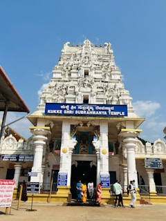 A temple with a richly carved gopura featuring intricate sculptures stands under a clear blue sky. The entrance is adorned with columns, and a group of people, some in traditional attire, are entering the temple.