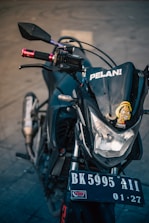 A close-up view of the front of a motorcycle, focusing on the headlight area with a cartoon sticker and the word 'PELAN!' on the windshield. The motorcycle's handlebars are visible with red grips, and the license plate reads 'BK 5995 AII 01-27'. The background is a neutral, blurred concrete surface.