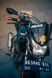 A close-up view of the front of a motorcycle, focusing on the headlight area with a cartoon sticker and the word 'PELAN!' on the windshield. The motorcycle's handlebars are visible with red grips, and the license plate reads 'BK 5995 AII 01-27'. The background is a neutral, blurred concrete surface.