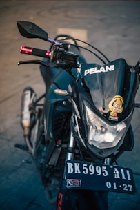A close-up view of the front of a motorcycle, focusing on the headlight area with a cartoon sticker and the word 'PELAN!' on the windshield. The motorcycle's handlebars are visible with red grips, and the license plate reads 'BK 5995 AII 01-27'. The background is a neutral, blurred concrete surface.