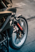 A close-up view of a motorcycle showing parts of the front wheel, fork, and fender. There are various stickers on the bike, including brands like Ipone, and a Michelin Man logo. The wheel is detailed with a brake disc and caliper, and there is a visible red cable.
