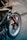 A close-up view of a motorcycle showing parts of the front wheel, fork, and fender. There are various stickers on the bike, including brands like Ipone, and a Michelin Man logo. The wheel is detailed with a brake disc and caliper, and there is a visible red cable.