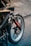 A close-up view of a motorcycle showing parts of the front wheel, fork, and fender. There are various stickers on the bike, including brands like Ipone, and a Michelin Man logo. The wheel is detailed with a brake disc and caliper, and there is a visible red cable.