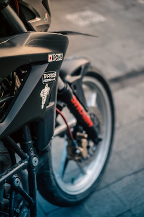 A close-up view of a motorcycle showing parts of the front wheel, fork, and fender. There are various stickers on the bike, including brands like Ipone, and a Michelin Man logo. The wheel is detailed with a brake disc and caliper, and there is a visible red cable.