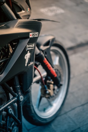 A close-up view of a motorcycle showing parts of the front wheel, fork, and fender. There are various stickers on the bike, including brands like Ipone, and a Michelin Man logo. The wheel is detailed with a brake disc and caliper, and there is a visible red cable.