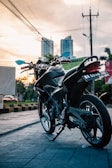 A sleek black motorcycle is parked on a paved urban street during sunset, with tall buildings visible in the background and lush greenery lining the area. The street is lined with yellow and black painted curbs, and there are streetlights and power lines overhead.