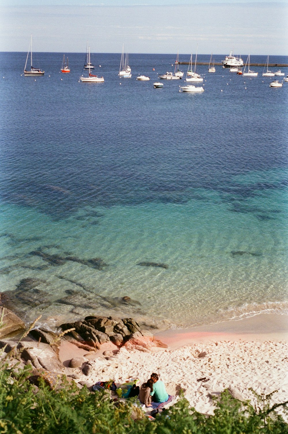 um grupo de pessoas sentadas em uma praia rochosa com barcos na água