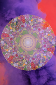 A vibrant mandala with intricate patterns, featuring a combination of floral and geometric designs. The mandala is filled with vivid colors, primarily purples, greens, and reds, and is set against a background of swirling purple and orange clouds, creating a mystical and dreamlike atmosphere.