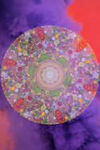 A vibrant mandala with intricate patterns, featuring a combination of floral and geometric designs. The mandala is filled with vivid colors, primarily purples, greens, and reds, and is set against a background of swirling purple and orange clouds, creating a mystical and dreamlike atmosphere.