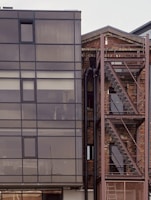 A modern building facade with a combination of glass and brick materials. The left side is dominated by large glass panels framed in dark metal, reflecting a muted sky. Adjacent to it, a brick structure features an industrial-style external staircase made of metal. The contrast between sleek modern glass and rustic brick gives an urban, architectural aesthetic.