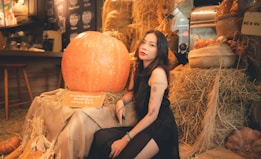 A large orange pumpkin is placed on a bale of hay, surrounded by rustic decor. A woman in a black dress with tattoos sits beside the pumpkin, resting her arm on it. The setting includes hay bales, baskets of produce, and warm ambient lighting, suggesting a cozy, farm-like environment.