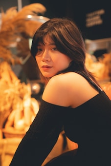 A person with long dark hair wearing a black off-shoulder top looks directly at the camera. The background is blurred with warm tones and decorative elements, possibly dried plants or wheat, creating an intimate and cozy atmosphere.