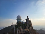 A large geodesic radar dome is situated atop a mountain peak surrounded by rocky outcrops and sparse vegetation. In the background, communication towers rise against a clear blue sky, with a view of distant hills.