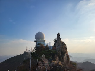 A large geodesic radar dome is situated atop a mountain peak surrounded by rocky outcrops and sparse vegetation. In the background, communication towers rise against a clear blue sky, with a view of distant hills.