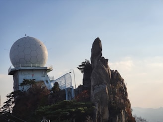 A geodesic dome structure, possibly a radar or observatory, is situated on a rocky cliff with sparse vegetation. The structure consists of a white, hexagonal-patterned sphere atop a building. Adjacent to the structure are natural rock formations, with some greenery scattered around. The background reveals a clear sky.
