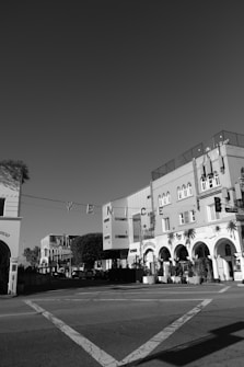 A black and white photograph captures an intersection in an urban area with distinctive architecture. Arched windows and doors characterize the buildings, and the word 'VENICE' is prominently displayed hanging across the street. Leafy trees and potted plants add a touch of greenery.