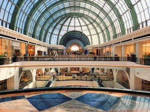 A spacious, modern shopping mall with an impressive glass dome ceiling, featuring multiple floors filled with retail stores. The area is bustling with people shopping and walking around.