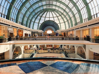 A spacious, modern shopping mall with an impressive glass dome ceiling, featuring multiple floors filled with retail stores. The area is bustling with people shopping and walking around.