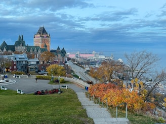A picturesque view of a historic cityscape featuring a prominent castle-like hotel with spires, surrounded by charming old buildings and autumnal trees. The foreground has a manicured garden and footpath leading down towards a wide promenade. A river and more urban structures expand into the distance under a partly cloudy sky.
