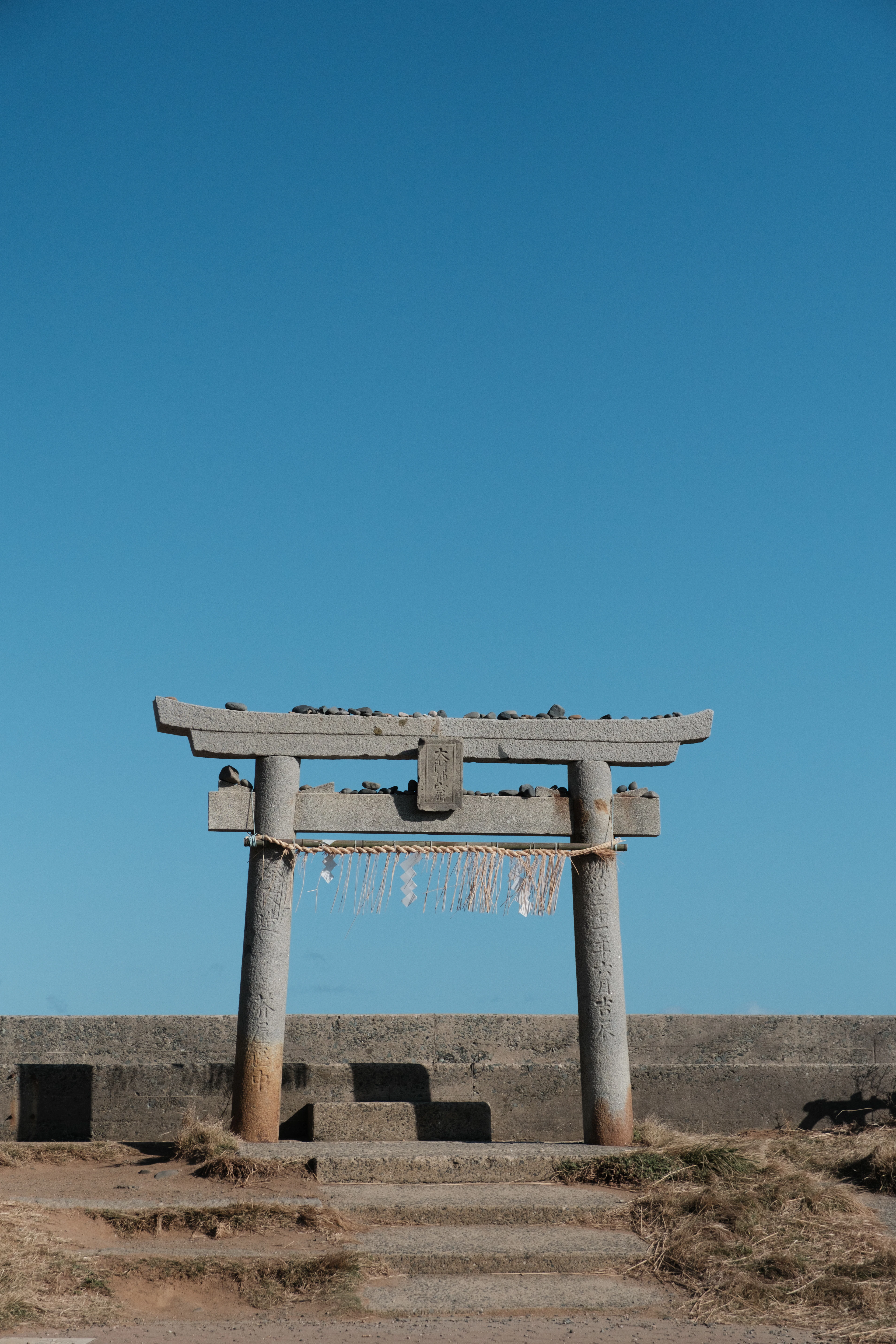 A stone torii gate stands alone against a clear blue sky on the coast.