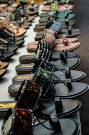 A display of various sandals and flip flops arranged neatly on a table. The sandals come in different styles, colors, and designs, including embellished straps and floral patterns. The foreground features dark sandals with intricate designs, while additional pairs are visible in the background.