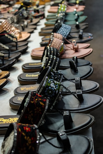 A display of various sandals and flip flops arranged neatly on a table. The sandals come in different styles, colors, and designs, including embellished straps and floral patterns. The foreground features dark sandals with intricate designs, while additional pairs are visible in the background.