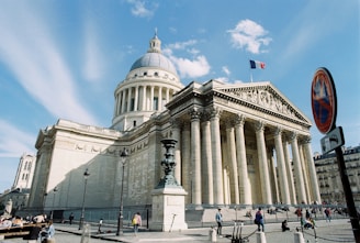 A neoclassical building with a large dome and detailed Corinthian columns is shown, topped with a French flag. The structure features classical stone architecture with intricate engravings and statues along the front pediment. In the foreground, there are people walking and sitting, and a no-parking sign is visible.