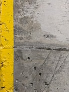 A close-up view of a concrete surface with a bright yellow painted line running vertically along the left side. The concrete appears textured with small holes and patches of discoloration, showcasing signs of wear and use.