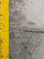 A close-up view of a concrete surface with a bright yellow painted line running vertically along the left side. The concrete appears textured with small holes and patches of discoloration, showcasing signs of wear and use.