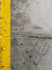 A close-up view of a concrete surface with a bright yellow painted line running vertically along the left side. The concrete appears textured with small holes and patches of discoloration, showcasing signs of wear and use.