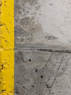 A close-up view of a concrete surface with a bright yellow painted line running vertically along the left side. The concrete appears textured with small holes and patches of discoloration, showcasing signs of wear and use.
